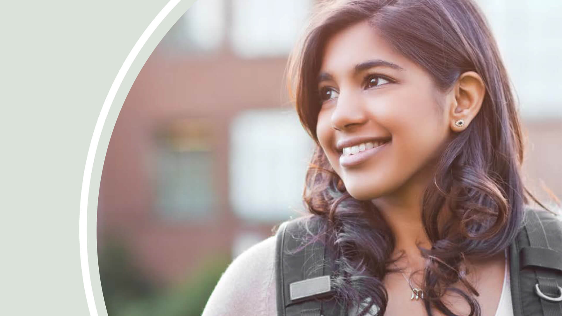 Young student smiling, carrying backpack. Buildings in background.