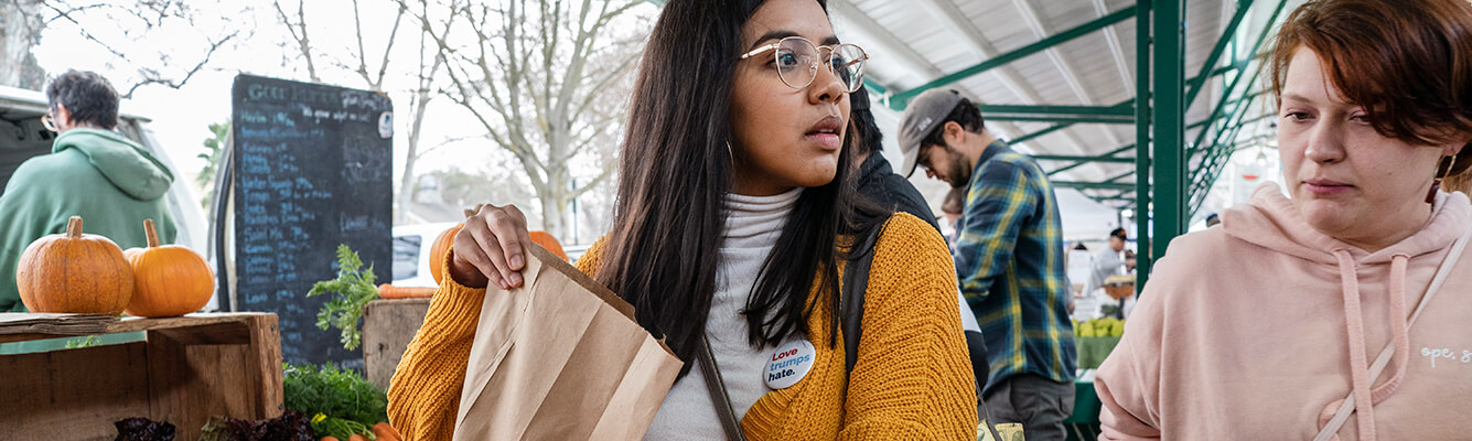 Karen Meza filling a paper bag at a farmers' market. A button on her yellow sweater reads Love Trumps Hate.