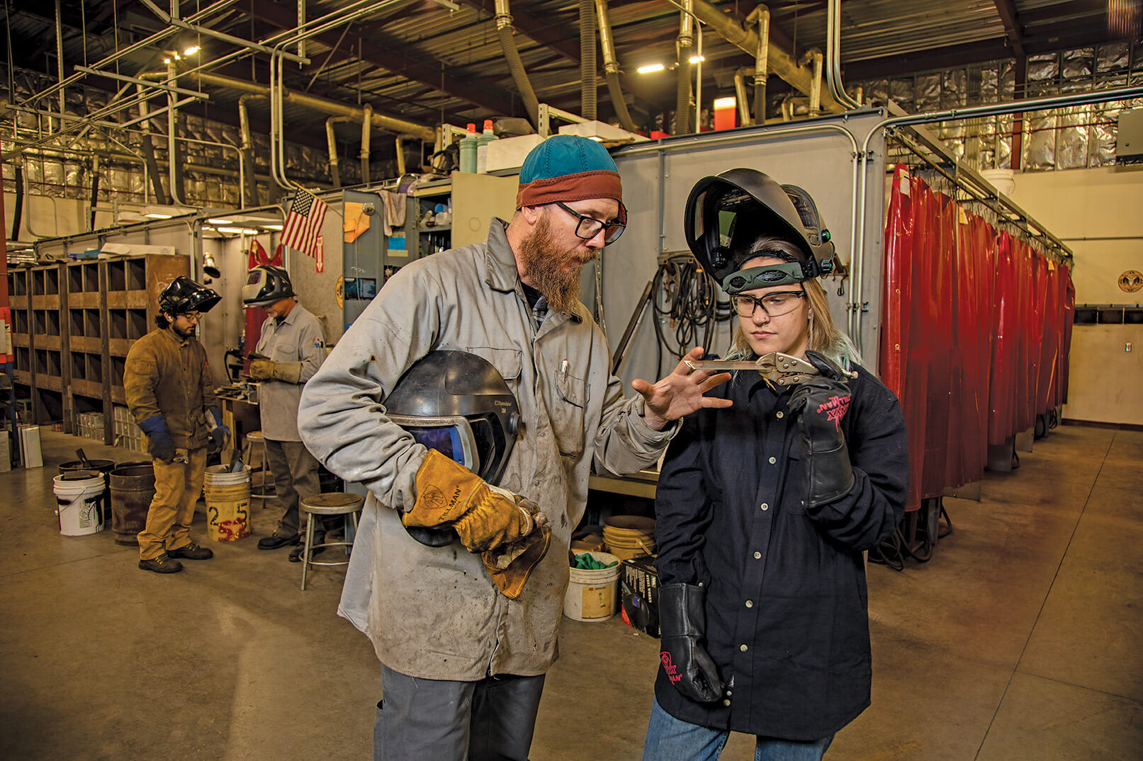 A woman in shop glasses and full protect welding suit clamps 2 samples of sheet metal between a pair of vice grips. A guy holding a helmet in one hand is placing his other hand on the metal.