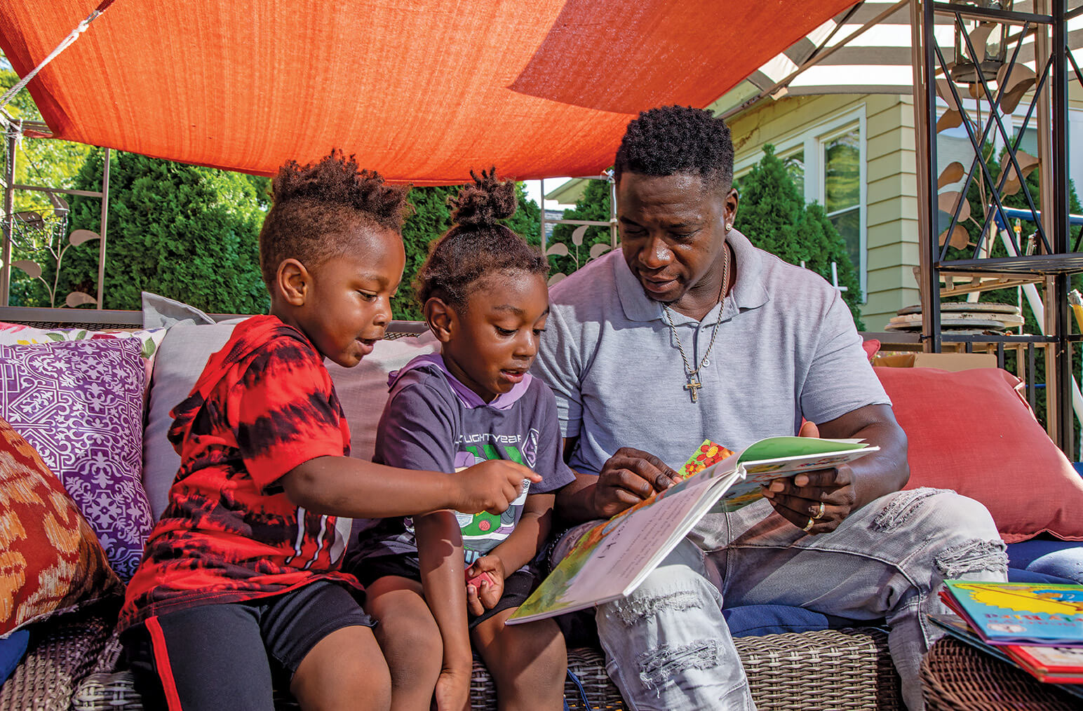 Kids listen to a story read by an adult male. They are seated outdoors.