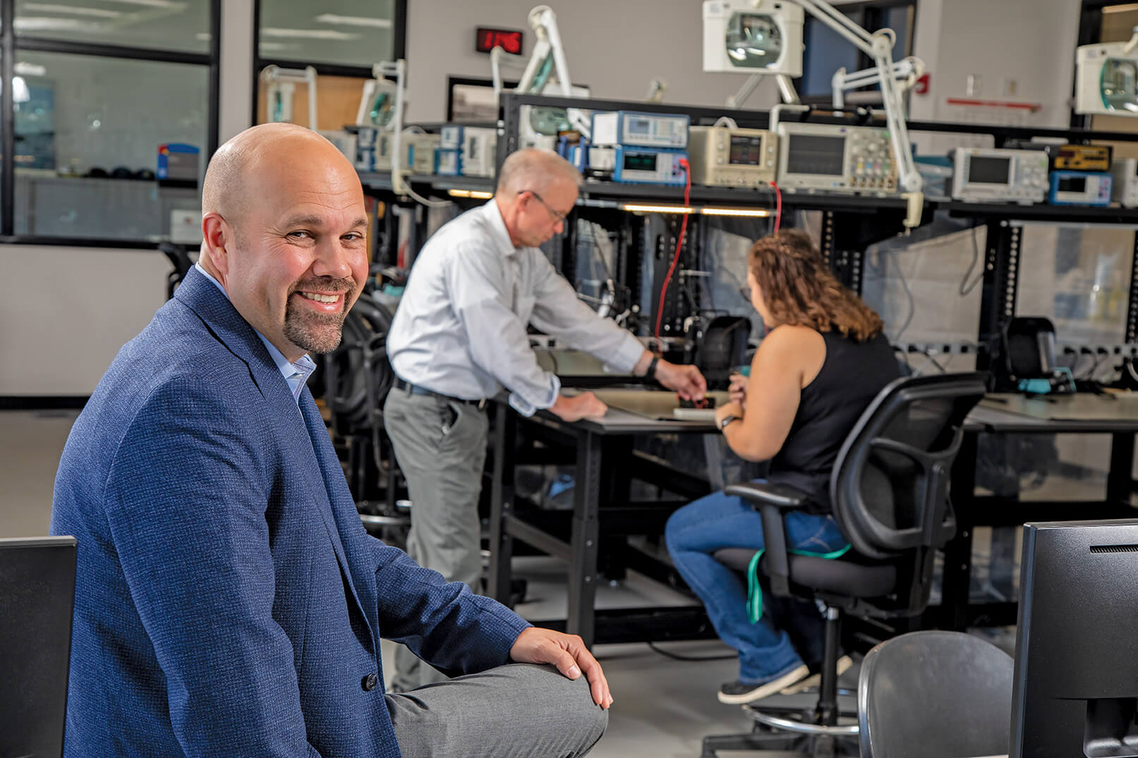 Man in blue blazer smiling. People in background at desk working. Lot of test equipment on shelves around them.