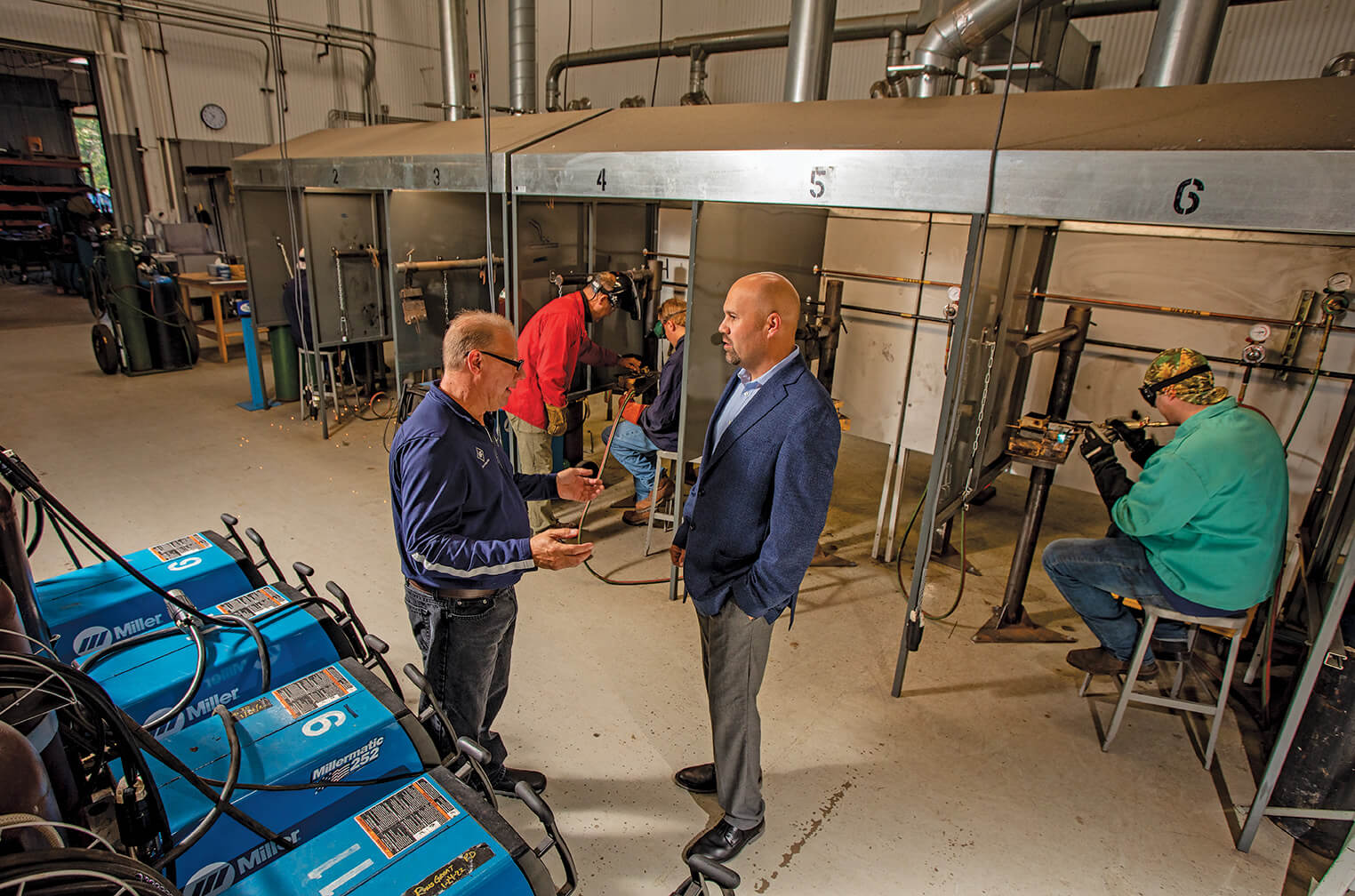 Overhead view of two men talking in an industrial environment with vent hoods under which 2 other men can be seen working in the background.