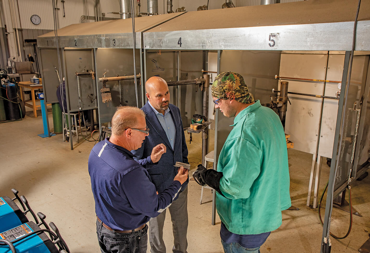 Posed shot of an older man explaining something to a younger man. The guy in the blue blazer from before is looking on. Welding stalls with ventilation hoods are in the background.