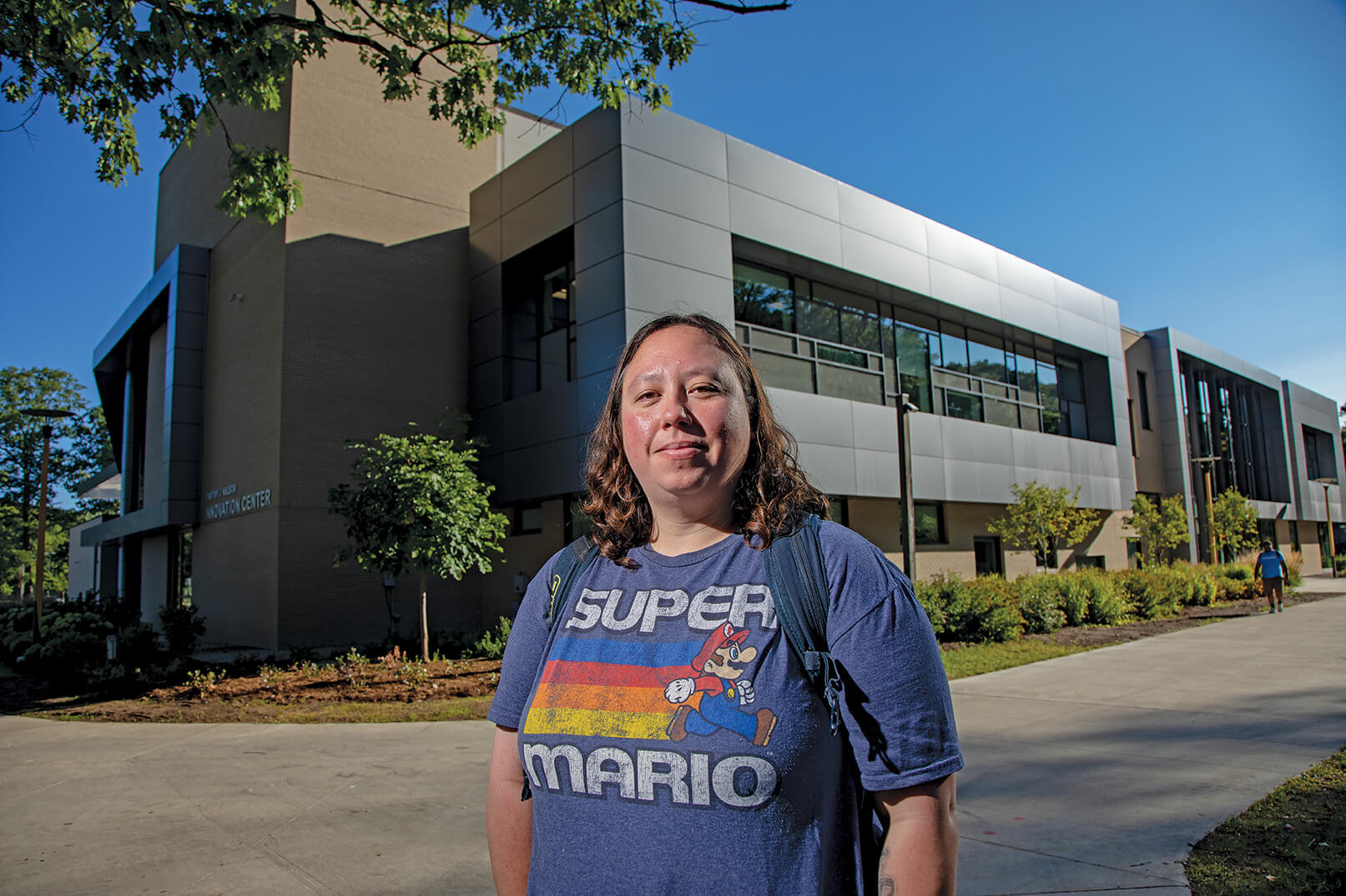 Woman with backpack, wearing a well-worn, blue Super Mario shirt. She's standing in front of a large industrial park building.