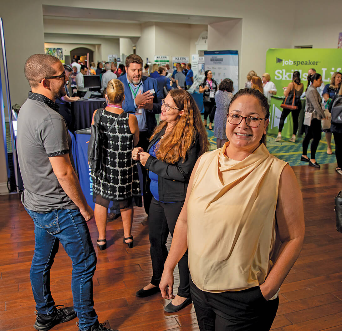 Woman with black-hair, pulled back, and glasses smiles. She is at a bustling jobs fair.