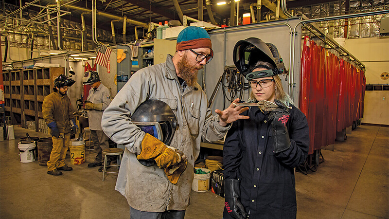 A woman in shop glasses and full protect welding suit clamps 2 samples of sheet metal between a pair of vice grips. A guy holding a helmet in one hand is placing his other hand on the metal.