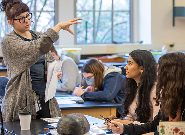 Earth science lab. A standing teacher gesticulating while explaining to students sitting at table.