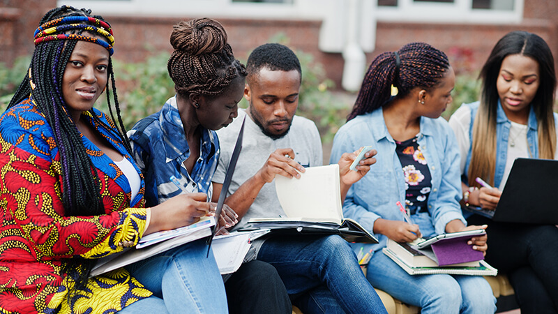 Group of five Black college students spending time together on campus at university yard. Black friends studying at bench with school items, laptops notebooks.